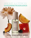 A bottle of OUAI Santorini fragrance mist centered on a white background, surrounded by peach, orange, grapefruit, blackberry, and a large pale peach flower. Text at the top reads 'BRIGHT & CITRUSY SANTORINI FRAGRANCE' and text at the bottom reads 'Notes of PEACH, HONEYSUCKLE, AMBER'.