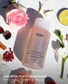 A bottle of OUAI Melrose Place body cleanser is shown in a styled flat-lay arrangement with a pink rose, white flowers, a lime slice, and berries on a white background with text overlay at the bottom.