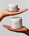Two hands holding OUAI body cream jars against a light gray background. The top jar is held by a lighter-skinned hand, and the bottom jar is held by a darker-skinned hand, showing two skin tones wearing the same product.