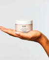 A hand holding a jar of OUAI Body Cream against a light gray background.