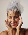 Woman with bubbles on her head and hands, smiling against a neutral background