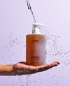 OUAI shampoo bottle being held with water being poured onto it against a light purple background