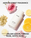 A white OUAI medium hair care formula bottle is displayed diagonally on a white background, surrounded by pink roses, sliced orange lemons, and white blossoms. Text overlays read 'MERCER STREET FRAGRANCE' at the top and 'Notes of ITALIAN LEMON, TURKISH ROSE, WHITE MUSK' at the bottom.