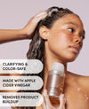 A woman with wet, foamy hair holds a bottle of OUAI Clarifying & Color-Safe Hair Cleanser against a soft mauve background, with text overlays highlighting key product benefits.