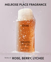 A bottle of OUAI Detox Shampoo centered against a pale pink background, with a stream of water pouring into the open bottle creating foam and bubbles. Large black text at the top reads 'MELROSE PLACE FRAGRANCE' and text at the bottom reads 'Notes of: ROSE, BERRY, LYCHEE'.