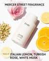 OUAI Mercer Street Fragrance Thick Hair Conditioner bottle in a white matte finish, styled in a flat lay with pink roses, sliced lemons, and dried rosebuds on a white background, with text detailing the scent notes.