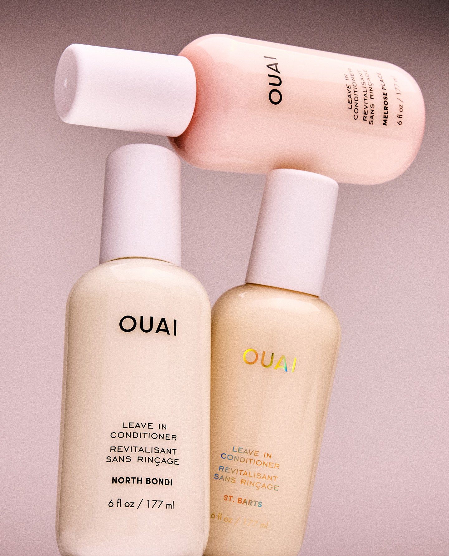Three OUAI leave-in conditioner bottles in soft pastel shades of cream, peach, and pale pink, arranged on a matching neutral background.