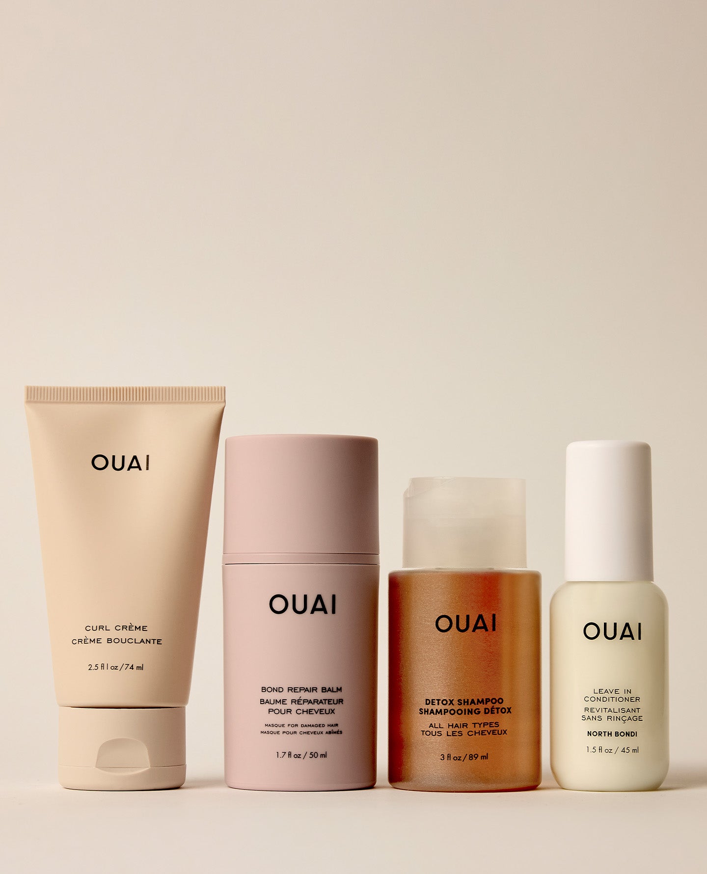Four OUAI skincare bottles and tubes lined up against a warm beige background. The set includes a beige cream tube, a dusty pink body butter bottle, an amber shampoo bottle, and a pale yellow whitening lotion bottle.