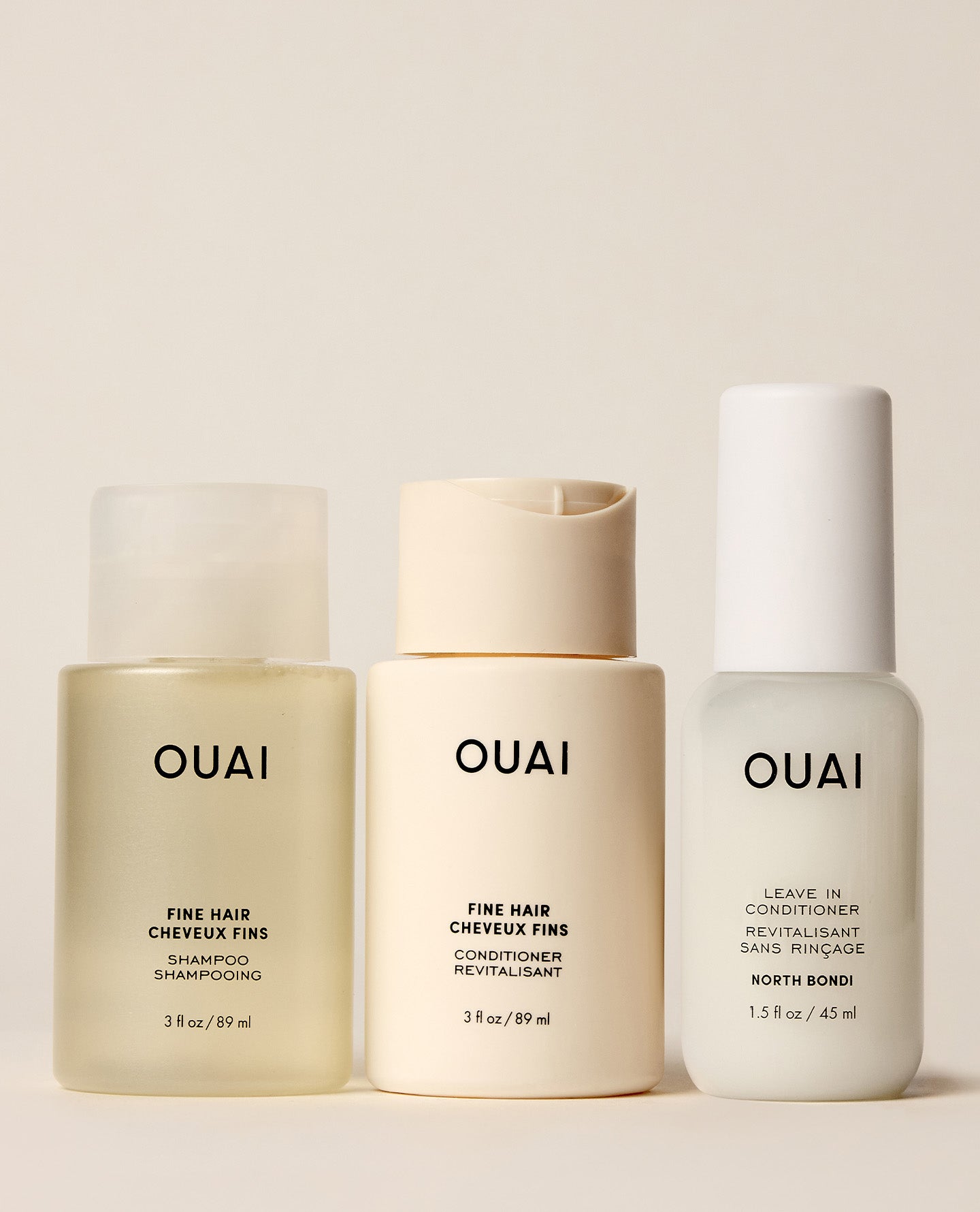 Three OUAI fine hair care bottles arranged side by side on a warm beige background: a translucent frosted bottle of shampoo, a cream-colored bottle of conditioner, and a white bottle of leave-in conditioner.