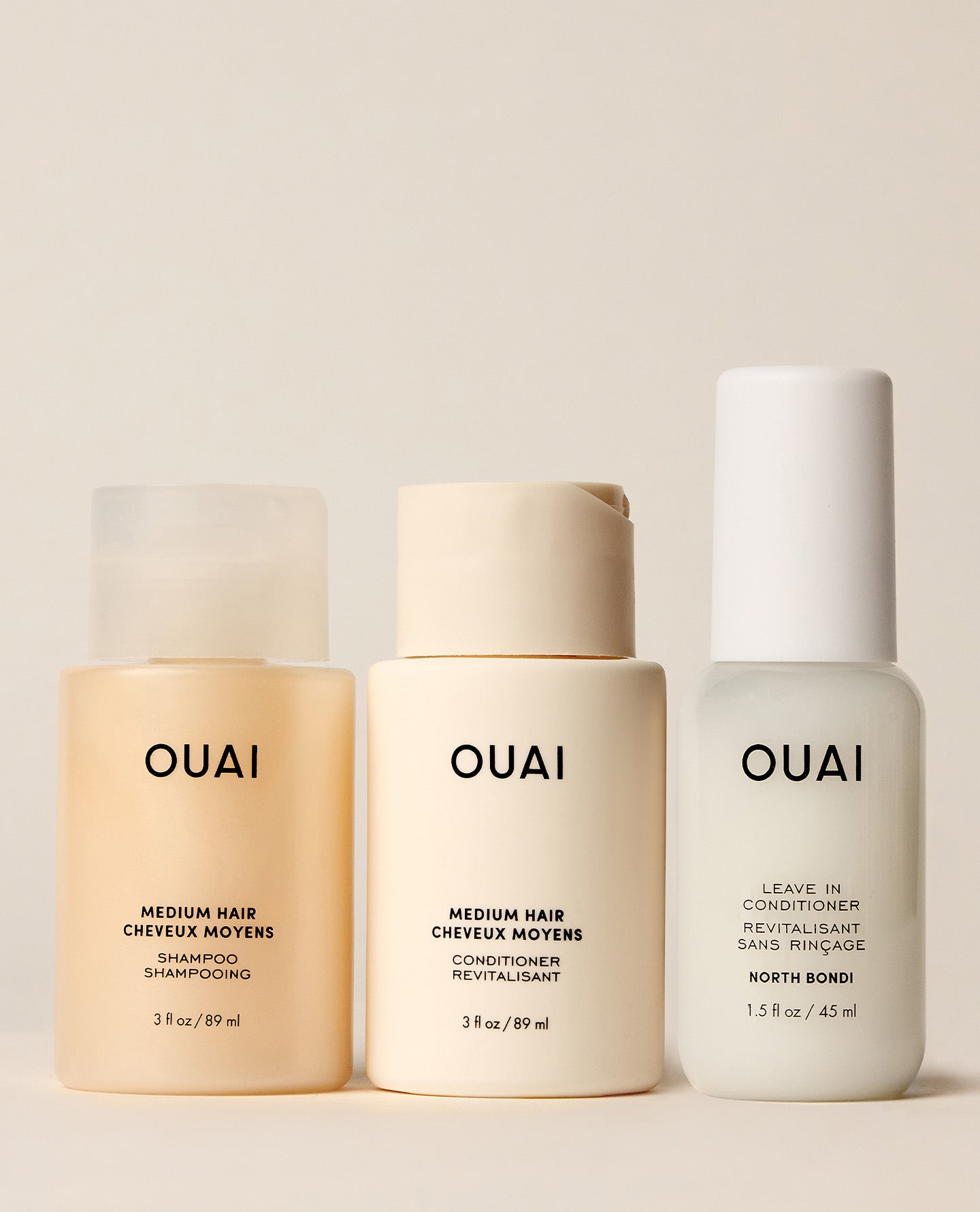 Three OUAI hair care bottles for medium hair arranged against a warm beige background: a translucent beige shampoo bottle, a solid beige conditioner bottle, and a white leave-in conditioner bottle with black text.