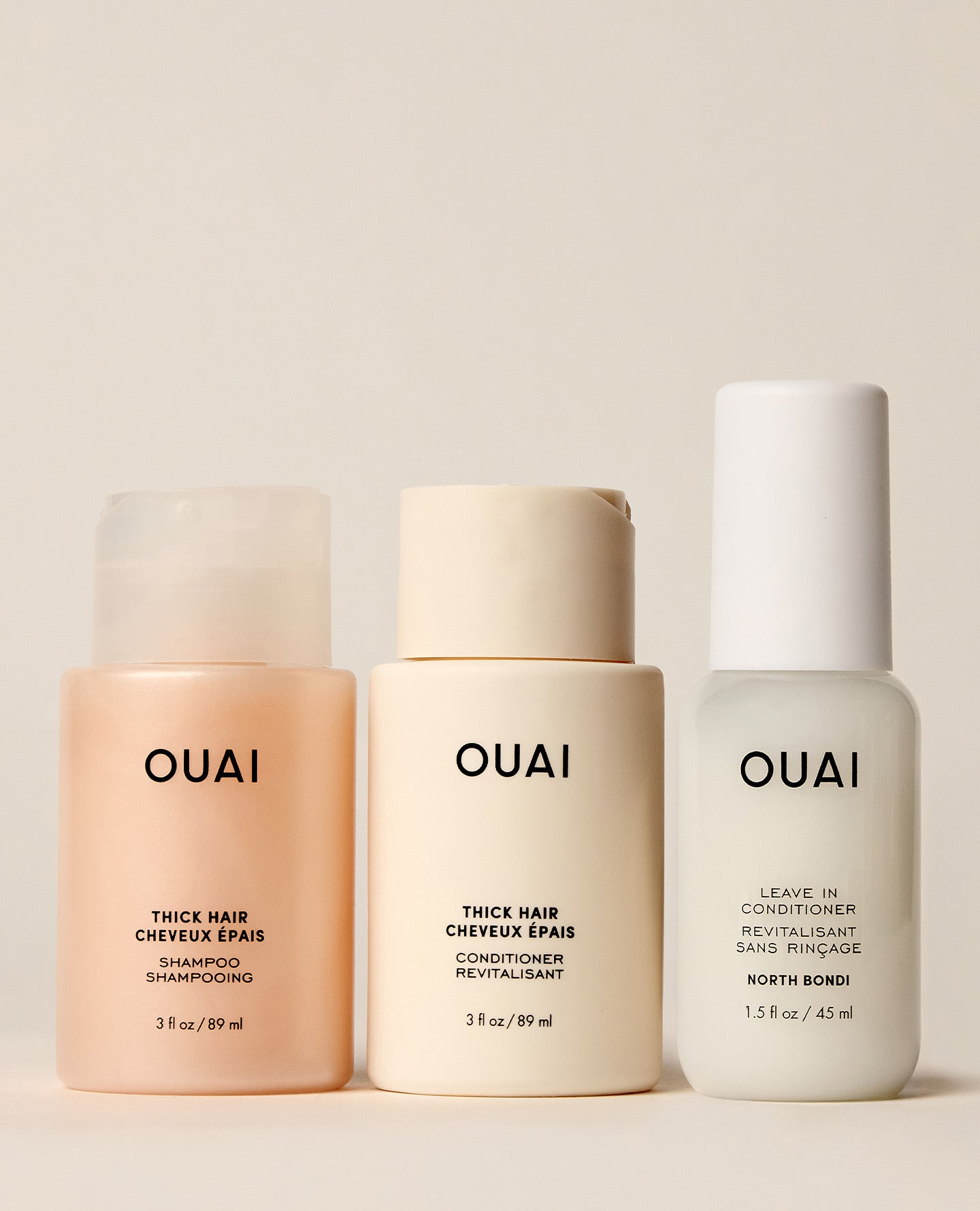 Three OUAI hair care bottles lined up against a warm beige background: a peach-colored shampoo bottle, a cream conditioner bottle, and a white leave-in conditioner bottle.