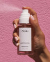 A hand with pale pink manicured nails holds a clear bottle of OUAI Fragrance Mist Melrose Place against a textured pink background.