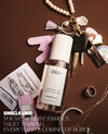 A perfume mist bottle labeled OUAI is arranged on a dark brown surface with lifestyle props including keys, a tassel, a pink key fob, a pearl necklace, a ring, and printed cards. Text at the bottom reads, 'SMELLS LIKE: YOU'RE ALMOST FAMOUS, VALET PARKING, EVERYTHING'S COMING UP ROSES.
