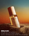 A bottle of OUAI body mist stands upright in sand against a warm sunset sky. The product has a pale pink cap and a cream label with black text.