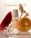 A bottle of OUAI St. Barts Fragrance centered on a reflective surface, surrounded by a sliced red dragon fruit, a sliced orange, white flowers, and amber droplets.