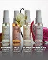 Four OUAI fragrance mist bottles arranged in a row on a reflective white surface, with flowers and fruit slices as styling props. Text labels below identify the scents: Santorini, Ibiza, St.