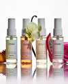 Four OUAI fragrance mist bottles arranged in a row on a reflective white surface, with white orchid flowers and fruit slices placed around them. The bottles contain translucent liquids in pale yellow, peach, pink, and lavender tones, with white spray tops and black text labels.