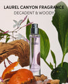 A clear cylindrical perfume bottle labeled 'OUAI' with lavender-toned liquid, centered among botanical props including green leaves, orange peels, wood bark, and a small green fruit. Large text at the top reads 'LAUREL CANYON FRAGRANCE' and 'DECADENT & WOODY'.