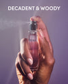 A hand holding a clear perfume bottle with pink liquid and a silver spray nozzle, with mist spraying from the atomizer. The background is a solid lavender-purple color, and the text 'DECADENT & WOODY' is written in white at the top.