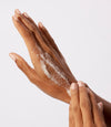 Hands applying white granular scrub product onto back of hand against white background.