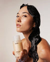 OUAI Medium Hair Shampoo held by woman with wet dark wavy hair.