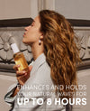 A woman with long wavy light brown hair holds a bottle of DUAL hair oil in front of a stone wall. The image features promotional text at the bottom: 'ENHANCES AND HOLDS YOUR NATURAL WAVES FOR UP TO 8 HOURS'.