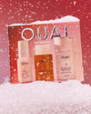 OUAI Hair Repair Kit set displayed in a snowy scene against a pink background. The set includes a peach-colored box with silver 'OUAI' lettering, three bottles and jars in soft peach and amber tones, and white faux snow in the foreground.
