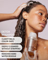 A woman with long dark curly hair lathered in shampoo holds a bottle of OUAI Detox Shampoo against a soft mauve background, with text overlays highlighting key benefits.