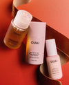 Three OUAI hair care products arranged on a warm orange background with a gold ribbon: a translucent bottle of leave-in shampoo, a tall box of bond repair balm, and a small bottle of leave-in conditioner.