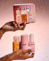 Two hands holding OUAI skincare bottles and a matching gift box, styled with white faux snow against a soft pink gradient background.