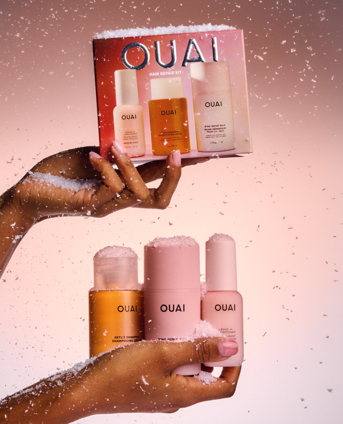 Two hands holding OUAI skincare bottles and a matching gift box, styled with white faux snow against a soft pink gradient background.