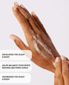 A close-up of a person's hand and wrist with white exfoliating scrub applied, accompanied by three text callouts describing benefits: exfoliates the scalp & body, helps balance natural bacteria levels, and nourishes the scalp & body.