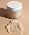 OUAI Scalp & Body Scrub in a frosted beige jar with a white lid, shown next to a textured smear of the sugar scrub on a warm beige background.