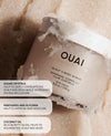 OUAI Scalp & Body Scrub in a frosted jar, partially embedded in sugar crystals on a light beige textured background, with three white text callout boxes on the left detailing ingredients.