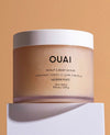 OUAI Scalp & Body Scrub in Melrose Place scent, shown in a frosted beige jar with a white lid, resting on a white pedestal against a warm beige background.