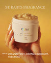 A hand holds an open white jar of OUAI St. Barts body scrub against a warm golden-yellow background.
