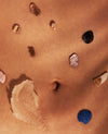 A close-up of warm tan skin with several small stones and shells embedded in it, including a blue seashell near the lower right and a pale shell near the upper left.