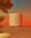 A jar of QUAI body and hair care product placed on sand, with a warm orange and peach gradient background and blurred natural elements around it.