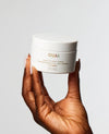 A hand with long manicured nails holds a white jar of OUAI Scalp & Body Scrub against a clean, light gray background.