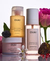 Three OUAI body care products arranged on a reflective white surface with floral props: a golden-yellow body cleanser bottle, a pale pink body crème jar, and a frosted clear fragrance mist bottle. Pink roses, a blackberry, white flowers, and small red berries surround the products.