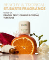 A fragrance bottle labeled CUAi centered on a white background, surrounded by a sliced orange, dragon fruit, white flowers, and green sprigs. Large text at the top reads 'BEACHY & TROPICAL ST.