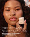 A close-up lifestyle shot of a woman holding a small white OUAI solid fragrance stick near her face, with text overlay reading 'TAKE ST. BARTS WHEREVER YOU GO' and 'BUILDABLE SOLID FRAGRANCE LASTS UP TO 8 HOURS'.