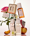 A styled product arrangement of OUAI fragrance mist bottles and packaging on a white background, surrounded by fruit slices (peach, dragon fruit, apple) and a pink rose.
