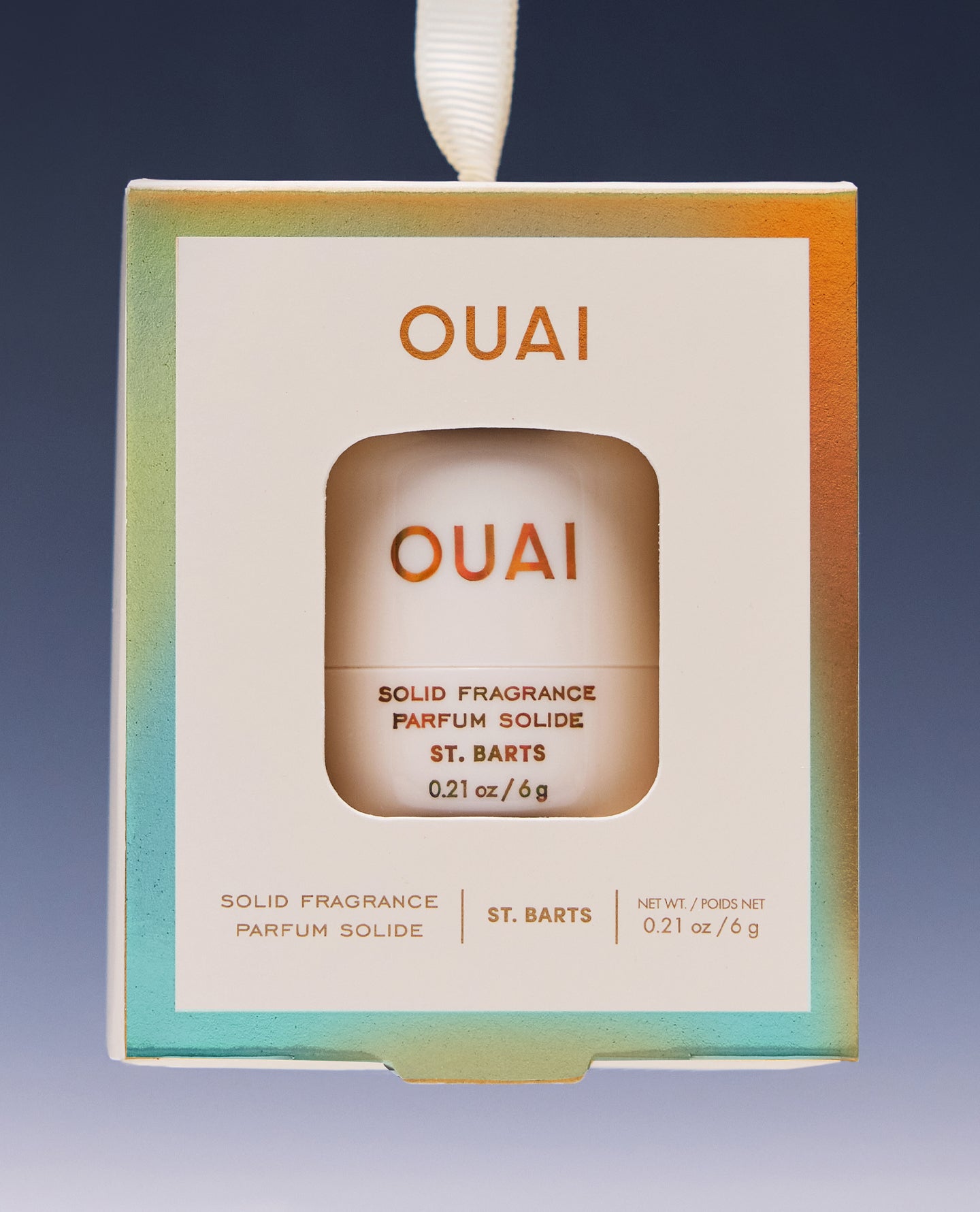 OUAI St. Barts solid fragrance perfume in a hanging box package with a white label and iridescent pastel border, shown against a dark blue gradient background.