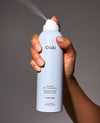 A hand holding a white spray bottle of OUAI Super Dry Shampoo against a solid gray background, with the spray mist visible.