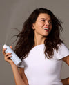 A woman with long, dark, windblown hair holds a white aerosol hair styling product in her hand, looking upward with a smile against a plain gray studio background.