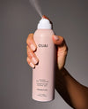 A hand holding a light pink aerosol can of OUAI Super Dry Shampoo against a dark gray background, with the spray nozzle emitting a fine mist.