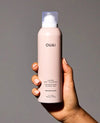 A hand holding a matte blush pink bottle of OUAI Super Dry Shampoo against a solid gray background.