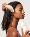 A woman with wet curly hair holds a white OUAI hair care bottle while using a light beige wooden comb on her hair, set against a clean light gray background.