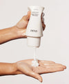 A hand holds a white OUAI Thick shampoo bottle dispensing thick white hair cream into another hand against a light gray background.